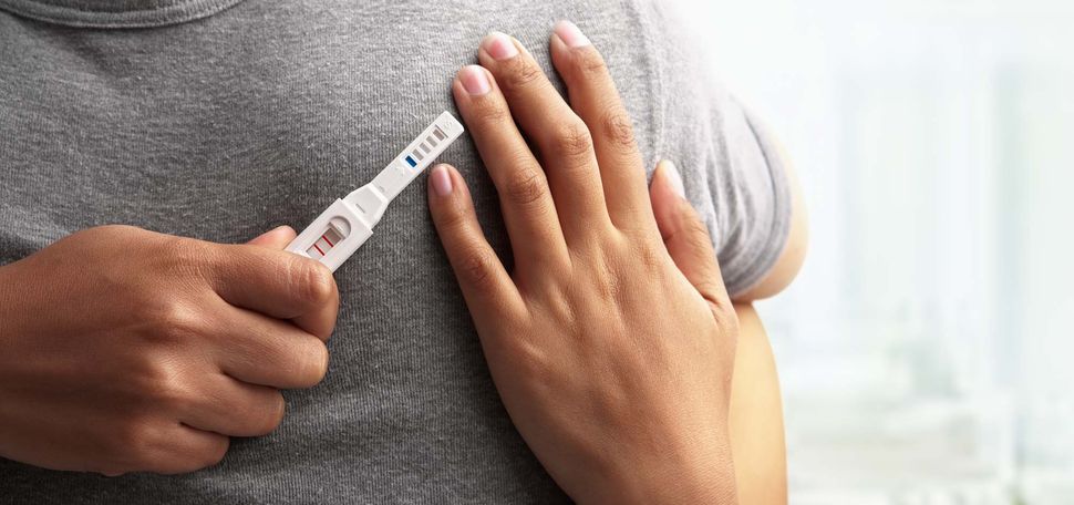 Photo of two partners, one dressed in a gray t-shirt, embracing as one holds a positive pregnancy test.