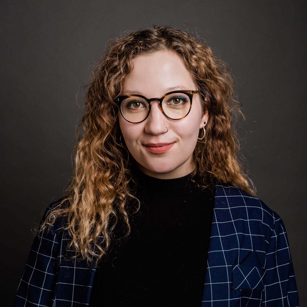 Photo of Allie Bouranova, a light skinned woman with blonde and brown curly hair. She smiles and wears glasses and a dark blue blazer with a light square pattern on it.