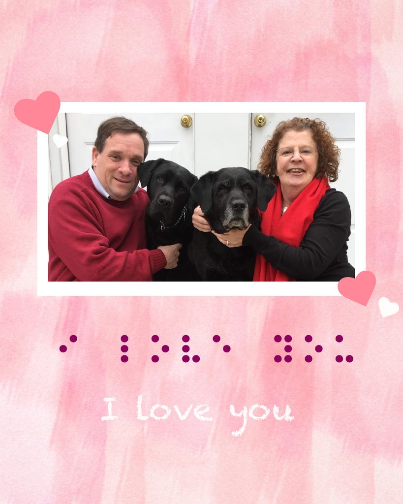 Photo of Megan Sullivan and her partner sitting together with two black labs; they both smile and hug the dogs. The photo is on a pink watercolor background and a few pink and white hearts are overlaid on the photo’s edges. Below the photo, the words “I love you” are written in both purple Braille and white text.