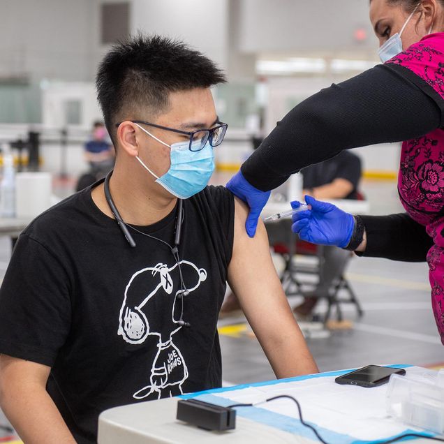 Photo of Yuqing Wu (COM’23), in a black Snoopy t-shirt, getting his vaccine from Kristin Lopes, registered nurse, in bright pink scrubs with a floral pattern, at the Moderna vaccine clinic at FitRec May 4. Both people wear masks.