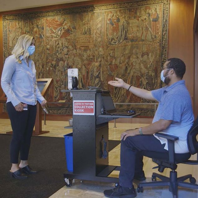 A male staff member at a COVID-19 Test Collection Kiosk prompts a woman to leave her testing swab vial in a tray on the kiosk table.