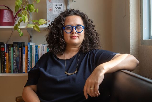 photo of Assistant professor Jessica Simes, a deeply tanned woman with curly dark brown hair and blue oval glasses, sits in her office and faces the camera. Her new book, Punishing Places, is seen on the bookshelf behind her.