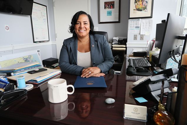 Photo of Rachael Rollins, a black women with freshly pressed hair, posing in her office in Boston. She wears a blue grey blazer over a white top as she smiles at her dark wooden desk.