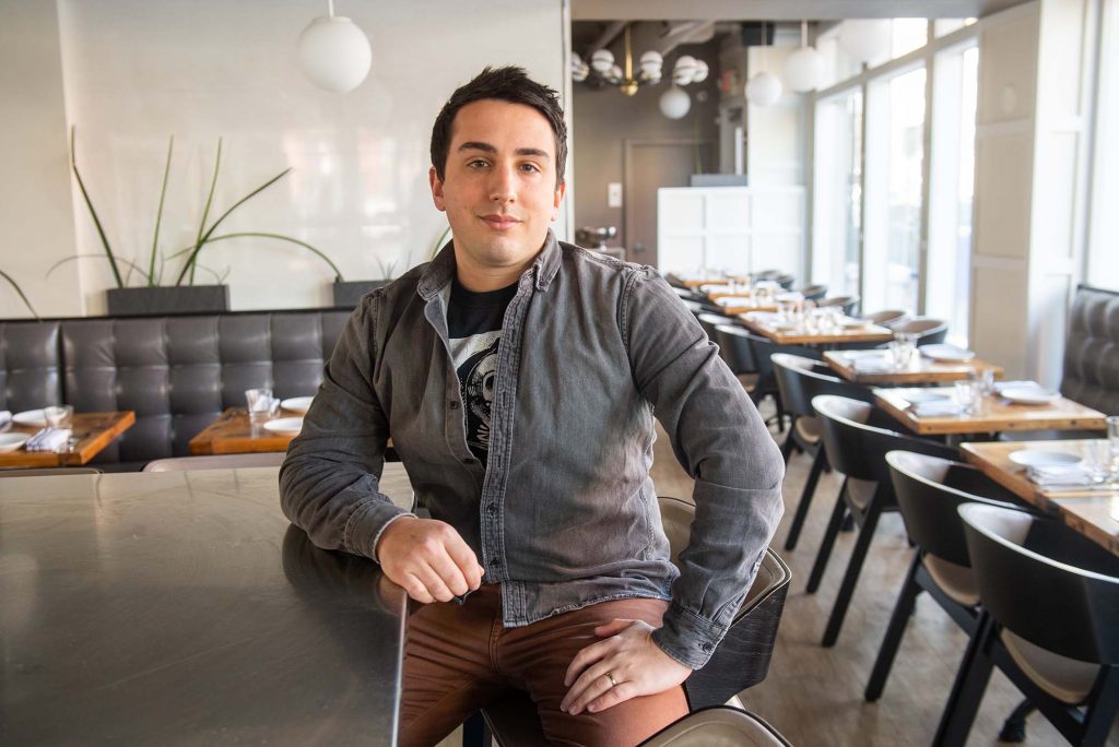 Photo of Seth Gerber, a young light-skinned man with short brown hair sitting at the bar of his South Boston restaurant Mida, wearing a washed gray jacket and brown pants. The wooden restaurant tables are set and have gray chairs or gray booth seats.