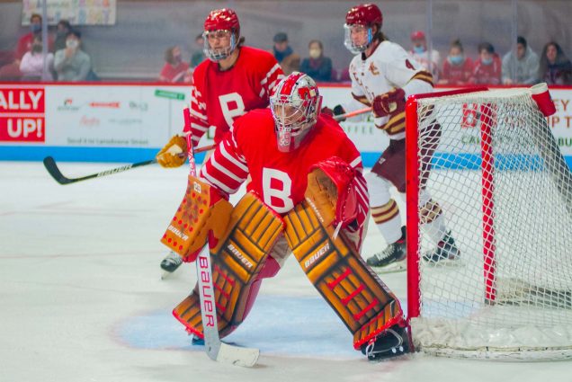 Photo of Drew Commesso (CAS’24) decked out in his hockey goalie attire. He wears a red BU jersey and stands at the ready in the side of the goal during a hockey game.