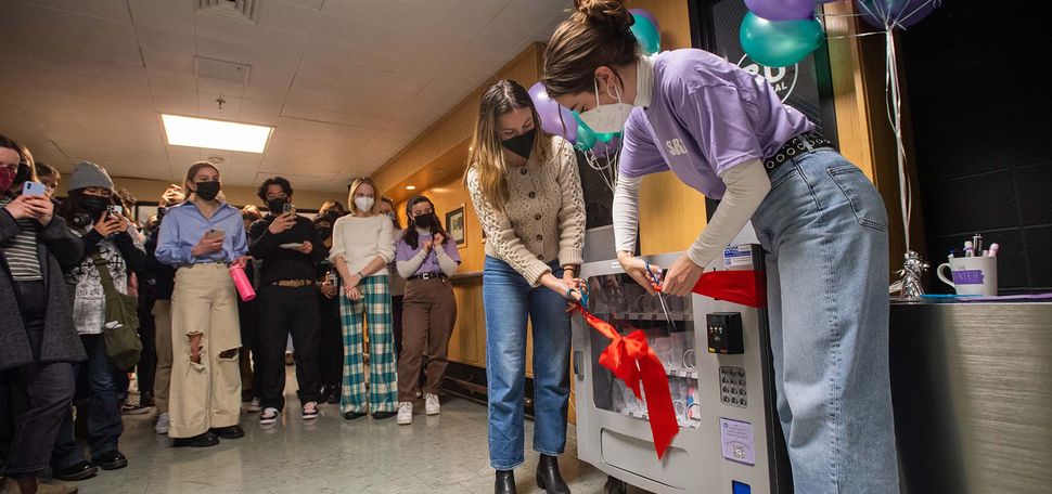 Photo of Students for Reproductive Freedom co-presidents Molly Baker (CAS’22), left, and Charlotte Beatty (CAS’22) cutting the ribbon on the new vending machine during a launch party in the GSU. Two masked students bend and cut a large red ribbon tied around the machine as masked onlookers circle around and clap.