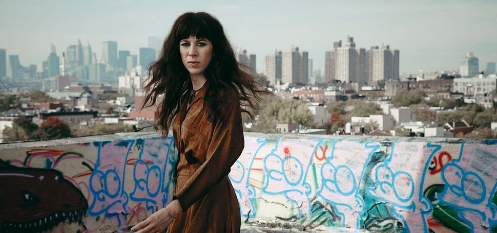 Photo of Missy Mazzoli, a white women with long brown hair. She wears a brown dress and is caught mid-turn on a roof with artistic graffiti covering it's walls. A city scene can be seen in the distance.