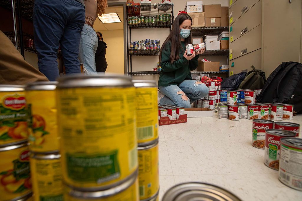 Photo taken behind a stack of yellow cans. In focus, in the corner of the room, is a young woman in a green sweatshirt and a mask who compares two cans in her hands.
