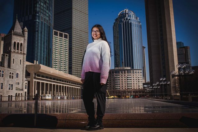 Photo of student Mira Dhakal, standing at the large reflection pool near the Christian Science Center. She smiles, wears a white and pink gradient sweater, and has long brown hair and glasses. The Boston city skyline and blue sky are seen behind her.