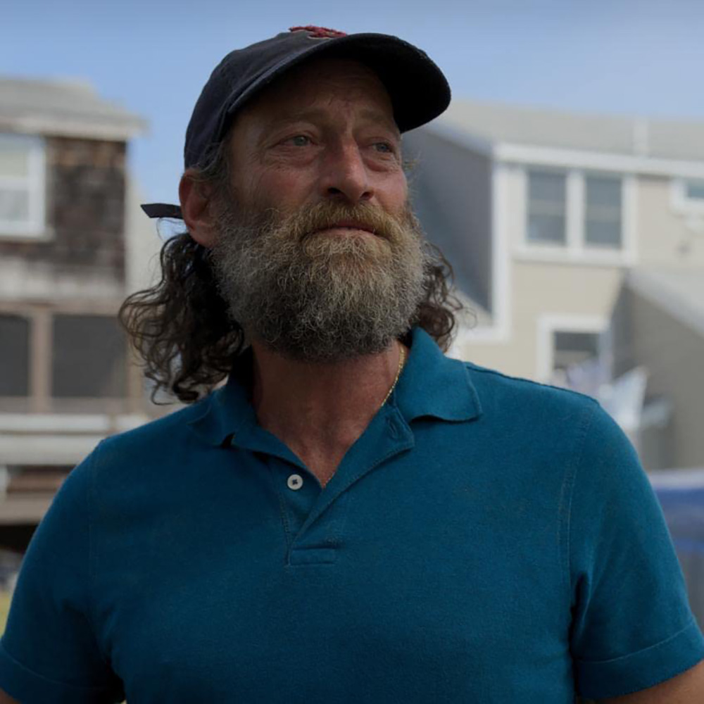 Photo of Troy Kotsur, playing a fisherman in the movie coda. He wears a blue polo shirt, a worn red sox baseball cap, and has a long, slightly unkempt graying beard and hair. Behind him, Gloucester houses are seen.