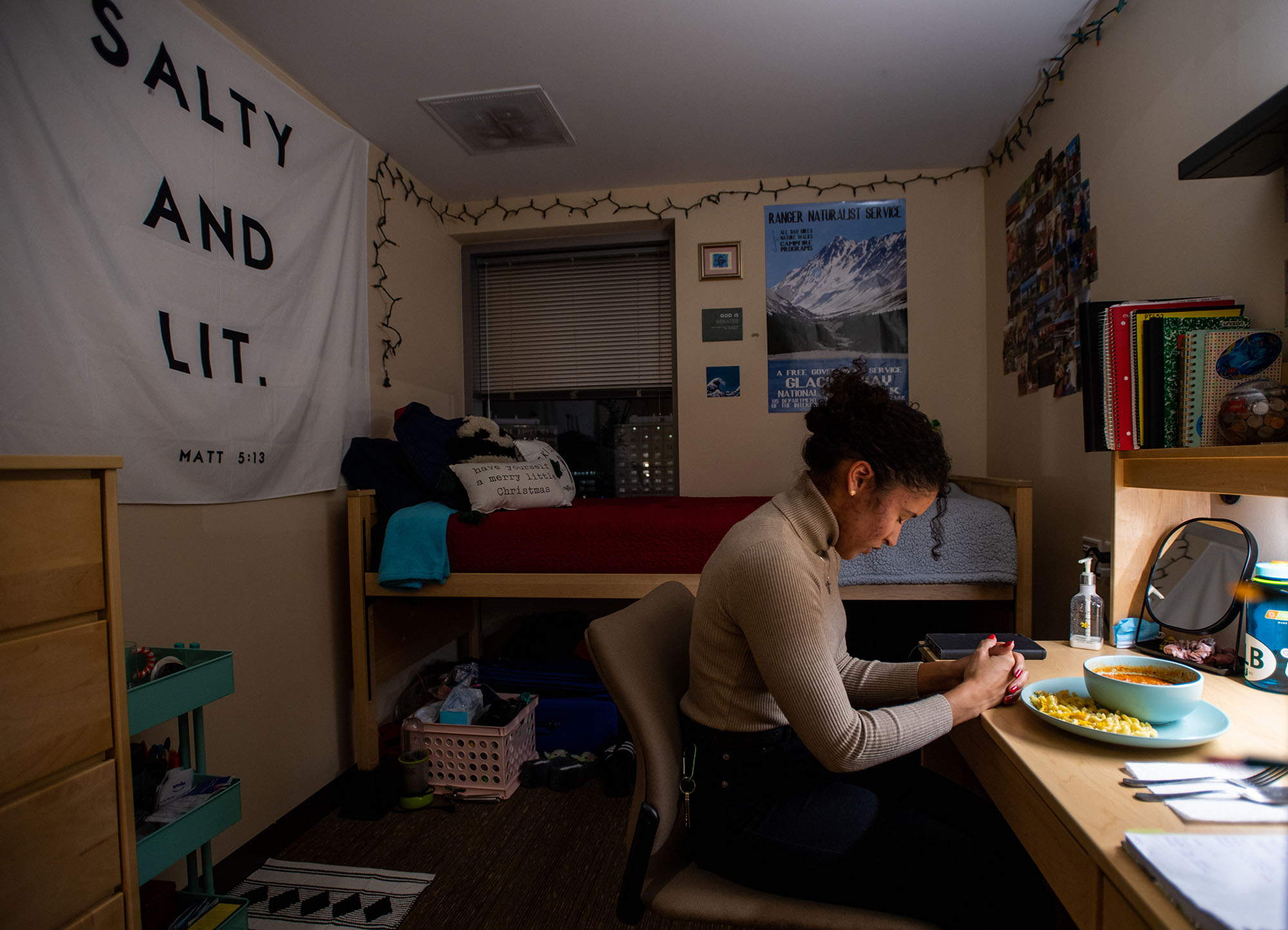 Emily Manz (SAR’23) says grace over her dinner in her Stuvi2 apartment. A tan young woman with black curly hair bows her head over her clasped hands as she sits at her desk in her dorm room.