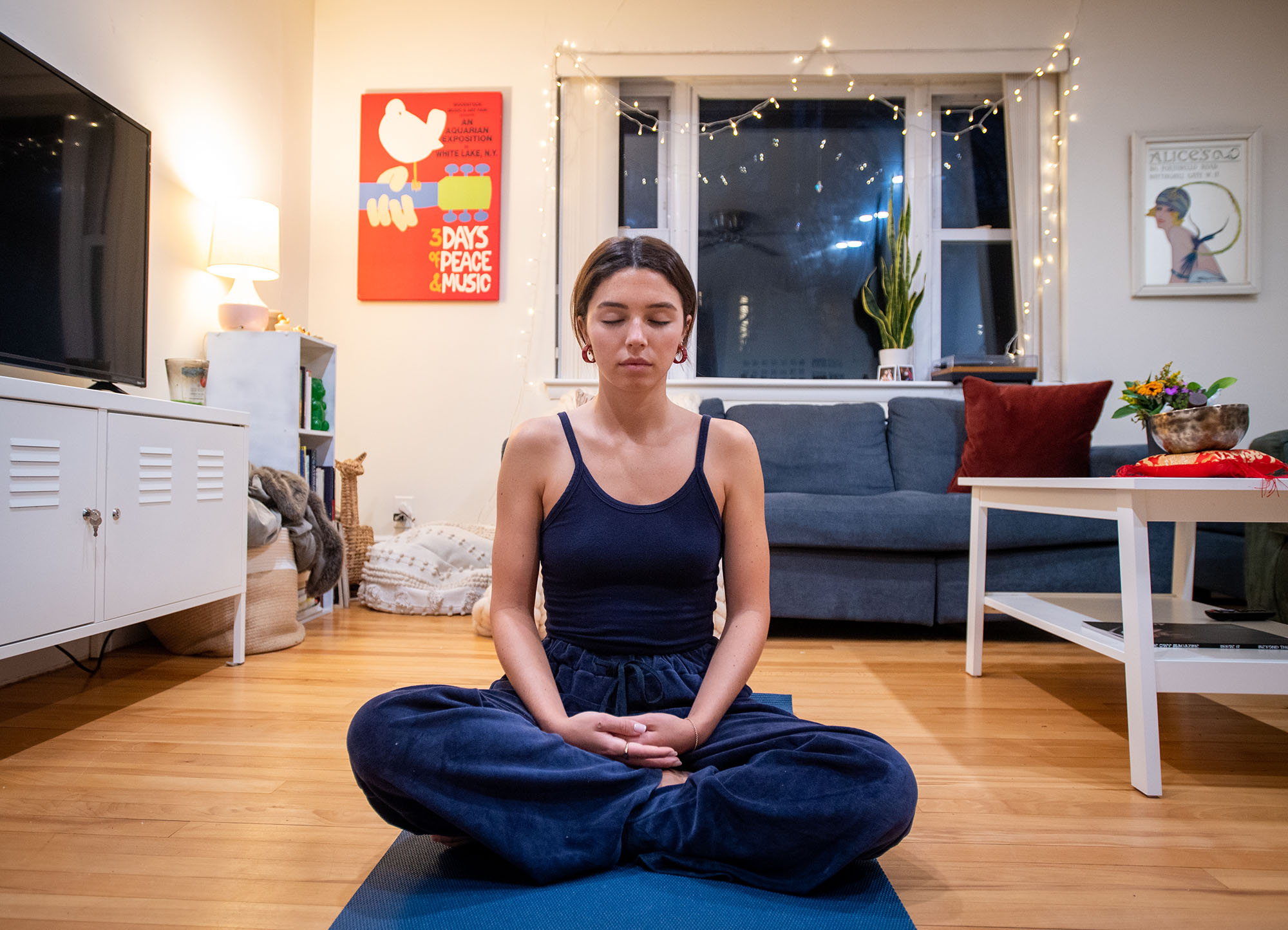 A photo of Aimee Mein (COM’22) meditating in her room. A white woman wearing a dark blue cami and pants sits with legs crossed and hands placed in her lap.