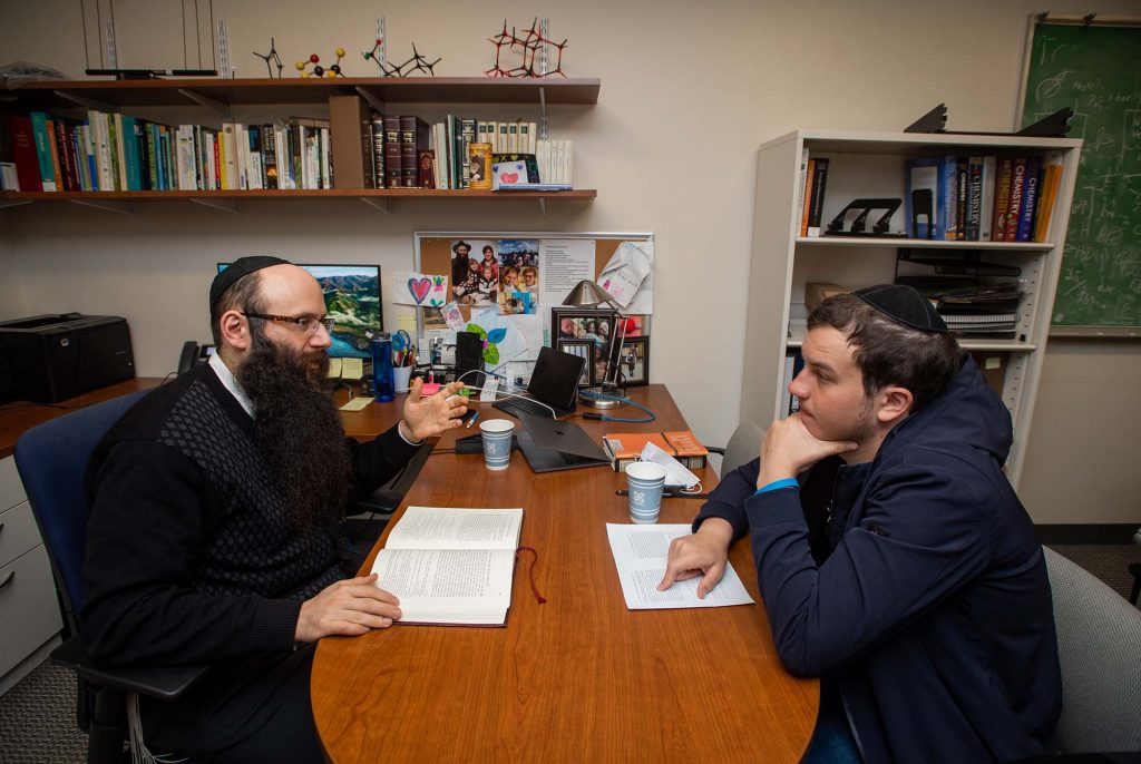 Photo of Rabbi Binyomin Abrams, left, learning the Torah with Rafael Kriger (CAS’22) in his Metcalf Science Center office. A Jewish man with a long beard and wearing a yarmulke sits on the other side of a desk and faces a younger Jewish man also wearing a yarmulke. The Torah sits between them