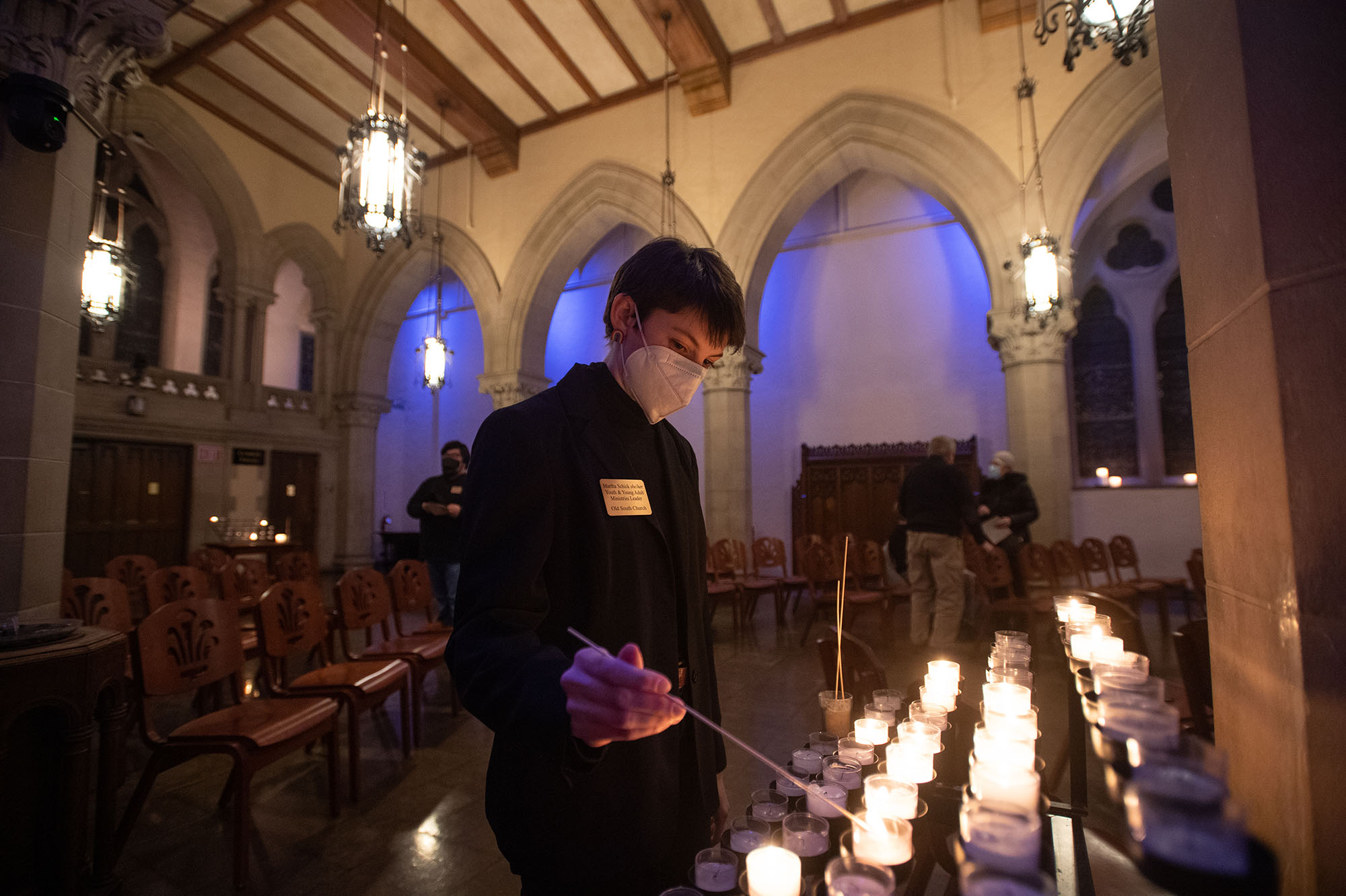 Photo of Martha Schick (MDiv’22) lighting a candle in Gordon Chapel. A white woman with short hair wearing a mask lights a candle with a long match in a darkened chapel