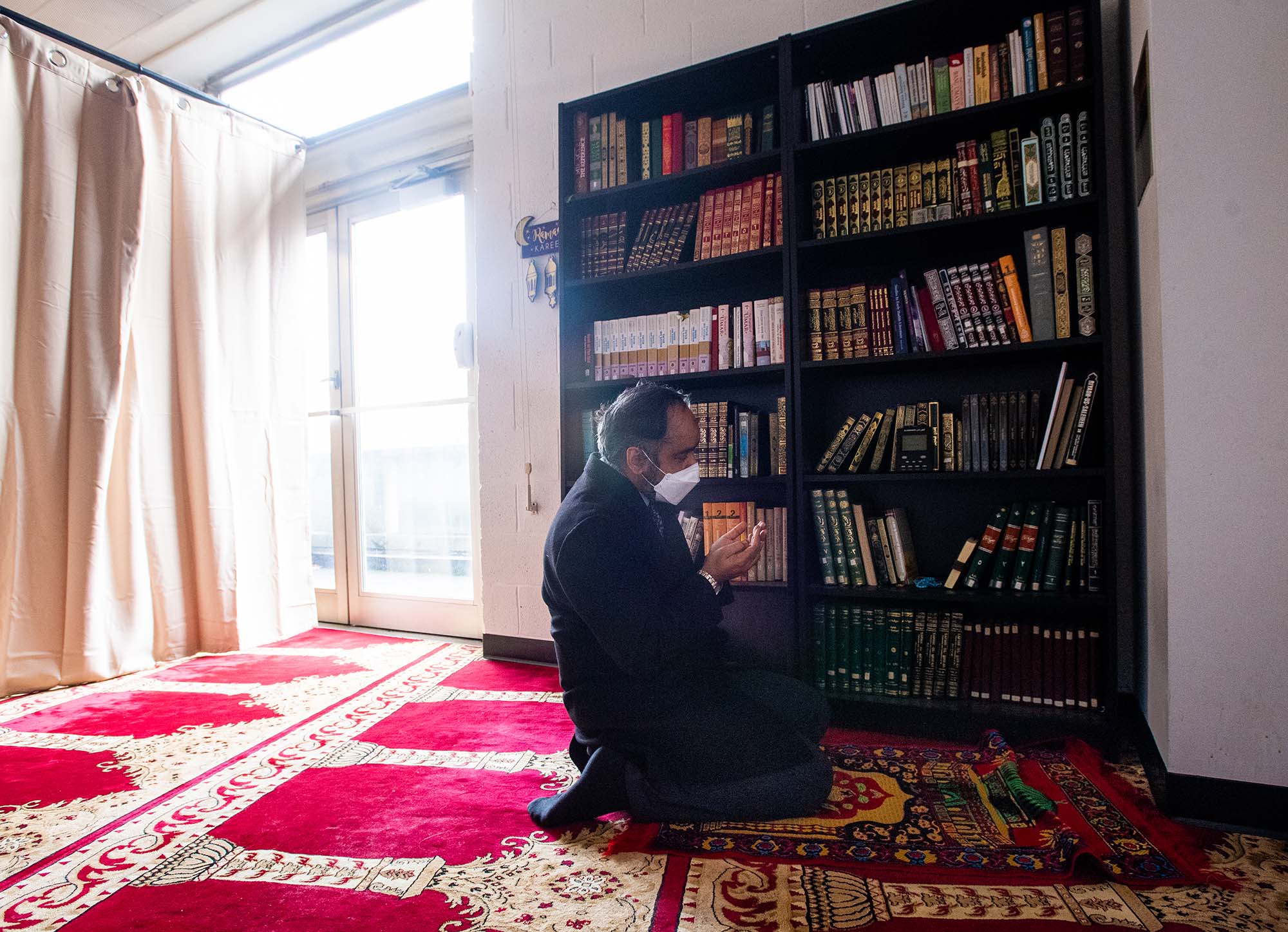 Photo of Professor Muhammad Zaman during Zuhr (noon) prayers at the ISBU prayer room in GSU. a man wearing a white mask kneels on an ornate rug with hands in prayer in front of him.