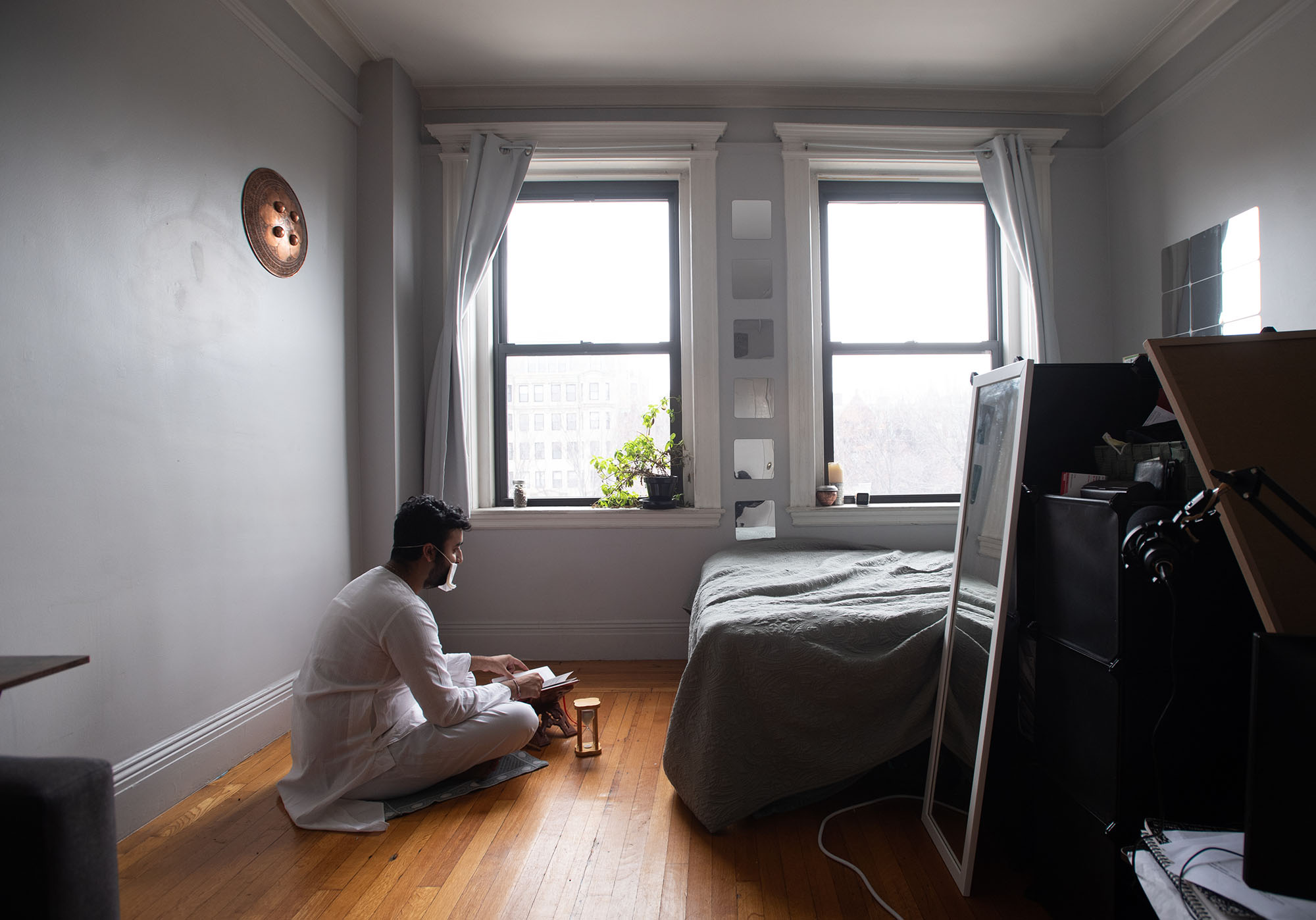 Photo of Adit Mehta, a tan man with black hair and beard, sitting cross-legged and wearing a white top and pants, on the floor in his room. He reads a book using the light from the window.