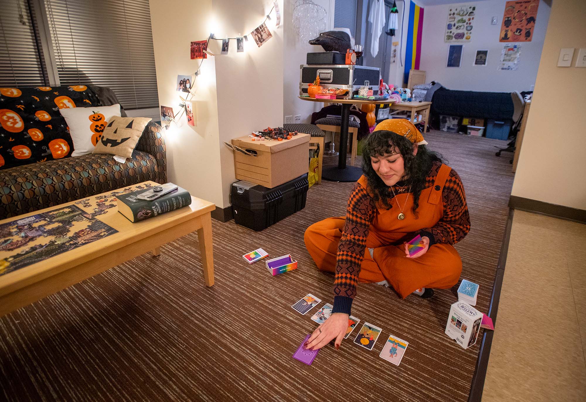 Photo of Zowie Rico (CAS’23), a white woman dressed in orange overalls, as she reads her Tarot and Prism Oracle cards in her Stuvi2 apartment