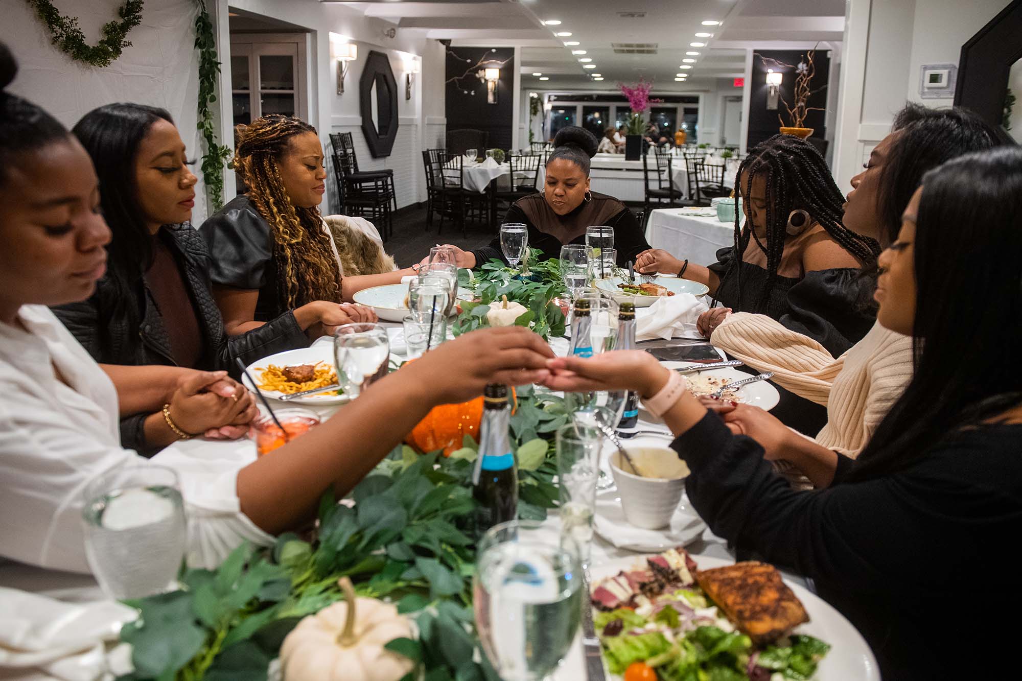Photo of 7 Black women seated and holding hands around a rectangular dining table with an assortment of food on it