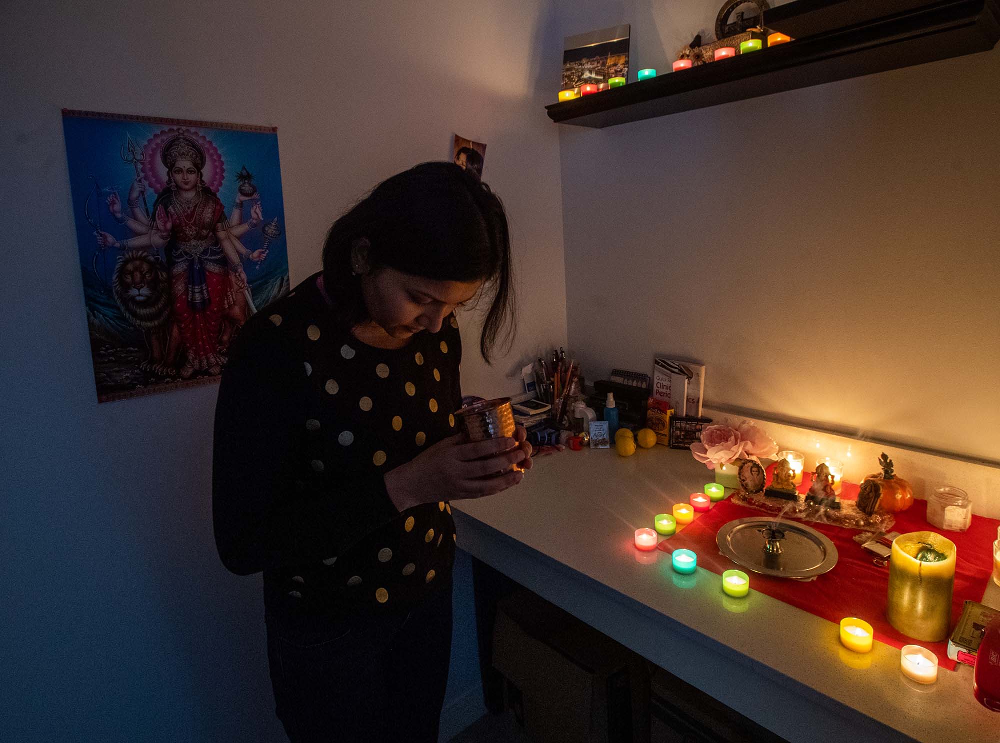Photo of Swati Gupta (GSDM’23), a brown woman with neck-length black hair, in her prayer/meditation space in her Boston home. She holds a cup made of copper and has head bowed as multi-colored candles are lit in the space.