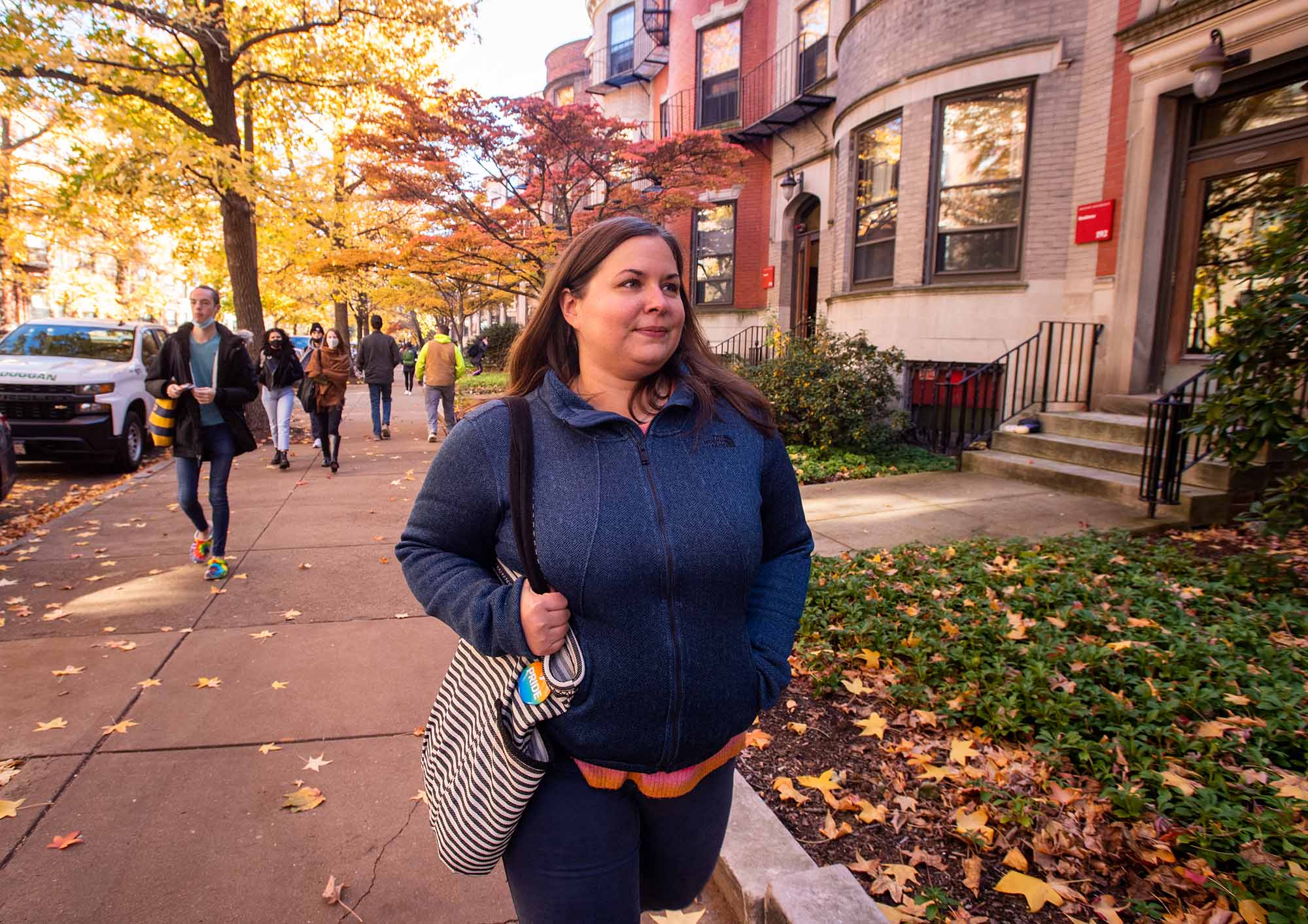 Photo of Rev. Kristen Hydinger, a white woman with brown hair and wearing a blue jacket, walking down a Boston street. Trees and leaves around her reflect Autumn in their color (yellow)