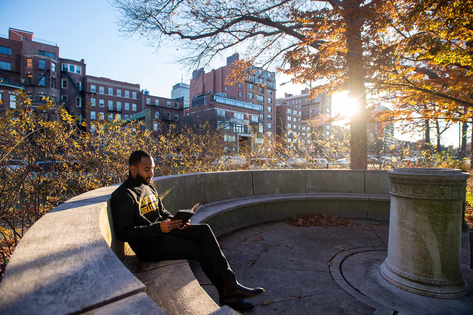 Photo of Interfaith leader Jonathan Allen sitting on a long stone bench along the Charles River. The sun can be seen peaking from behind the buildings in the background for a scenic photo.