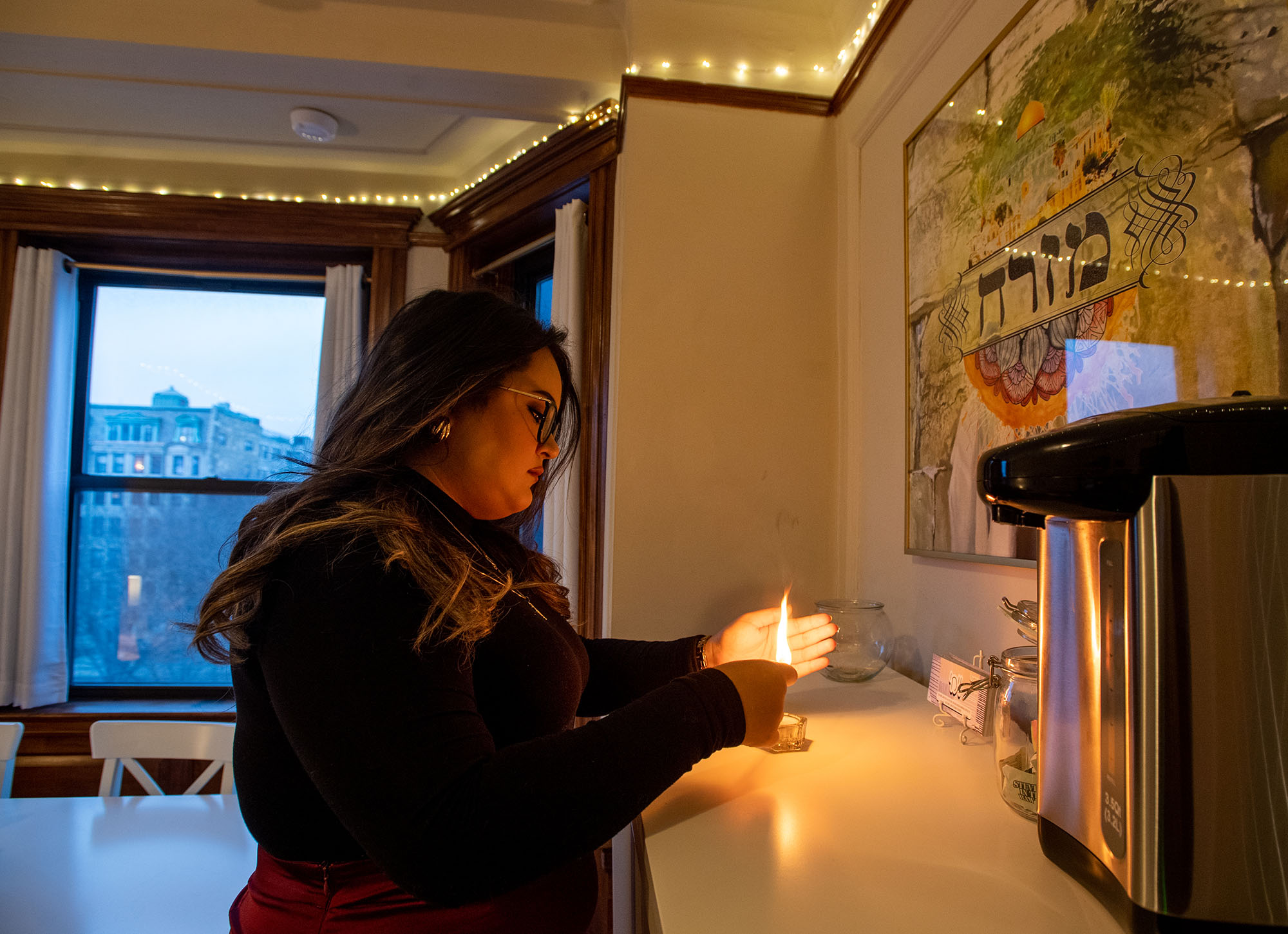 Photo of Kayla Marks (Pardee’23), a Jewish woman with long brown hair, demonstrating the lighting of one candle and the reciting of a blessing. She holds a lit match as she prepares for the lighting.