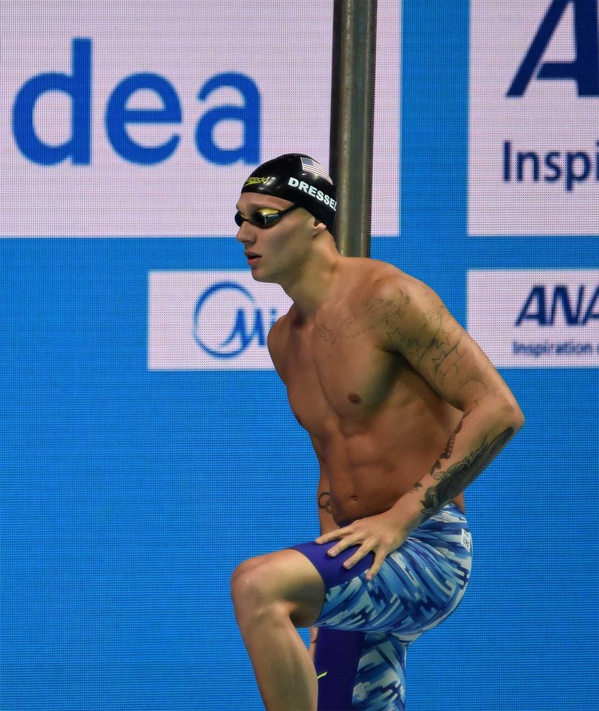 Photo of Swimmer Caeleb Dressel in a swimming cap and googles with his hand resting on his knee. He looks like he is at the edge of the pool about to swim.