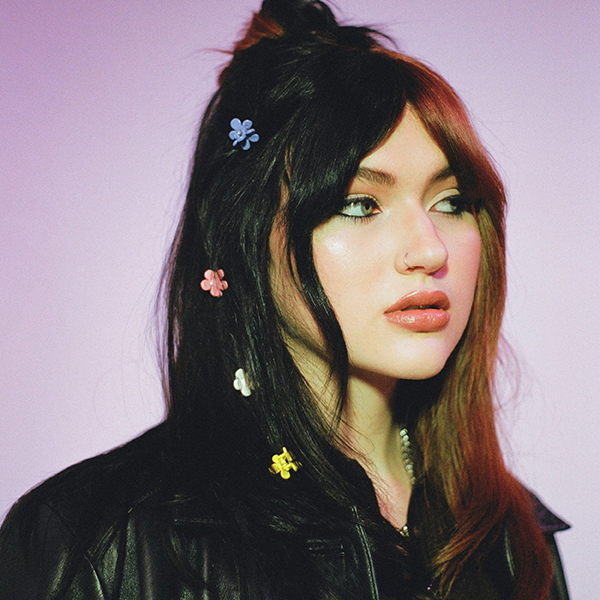 Photo of GAYLE, a white woman with black hair and a dyed brown bangs, looks to the right with an inquisitive expression. Small flower clips and sprinkled throughout her hair as she stands in front of a muted pinkish purple backdrop