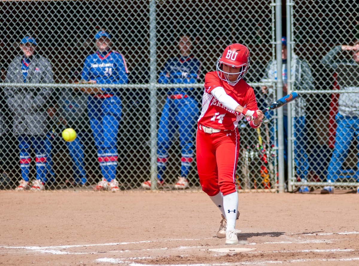 Photo of Jen Horita. A young women in a red BU softball uniform and helmet gets ready to swing at an oncoming softball at home plate. The opposing team in blue can be seen in the cage behind her.