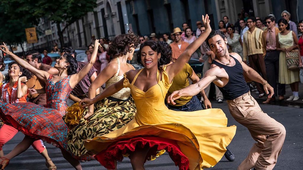 Photo of Ariana DeBose, center, in a gold dress with a red ruffly underskirt, during a number in West Side Story. Ariana DeBose, a young, brown-skinned woman with black hair pulled back into an up-do. Behind her, other dancers are mid-move, their arms and legs in the air. They dance at the center of a street with onlookers on the side walk.