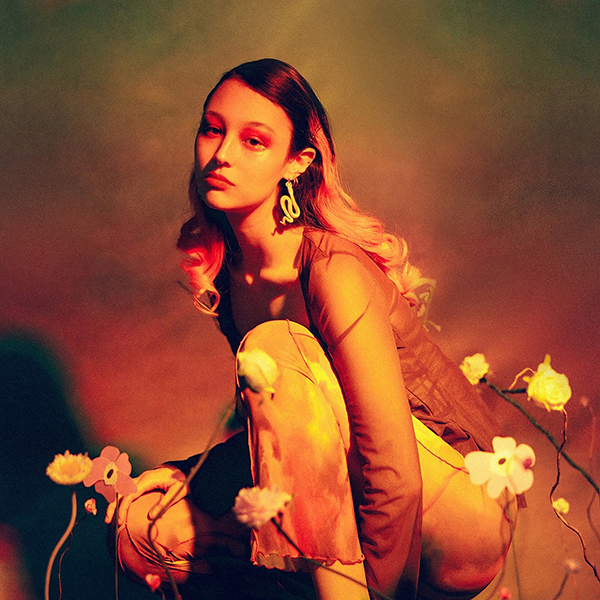 Album cover of Luna Li's album "Duality". Image shows Luna Li, squatting in front of a backdrop with various dried flowers in the foreground. Entire scene and image is tinted with dark oranges and green, giving off a mellow tone.
