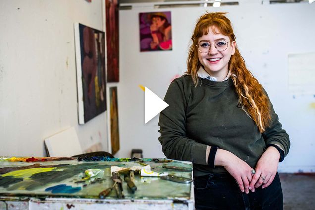 Photo of CFA graduate student, Eva Lewis, leaning against a white table covered with paint and paint mixing tools. She is a young white woman with long red hair and bangs, and wears round glasses and a green sweater. Behind her, other student's paintings hang on the wall. White video play button is overlaid.