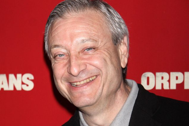 Candid photo of Fred Zollo as he stands in front of a red "Orphan" movie premiere background. An older white man with gray hair and wearing a grey shirt and black blazer smiles at the camera