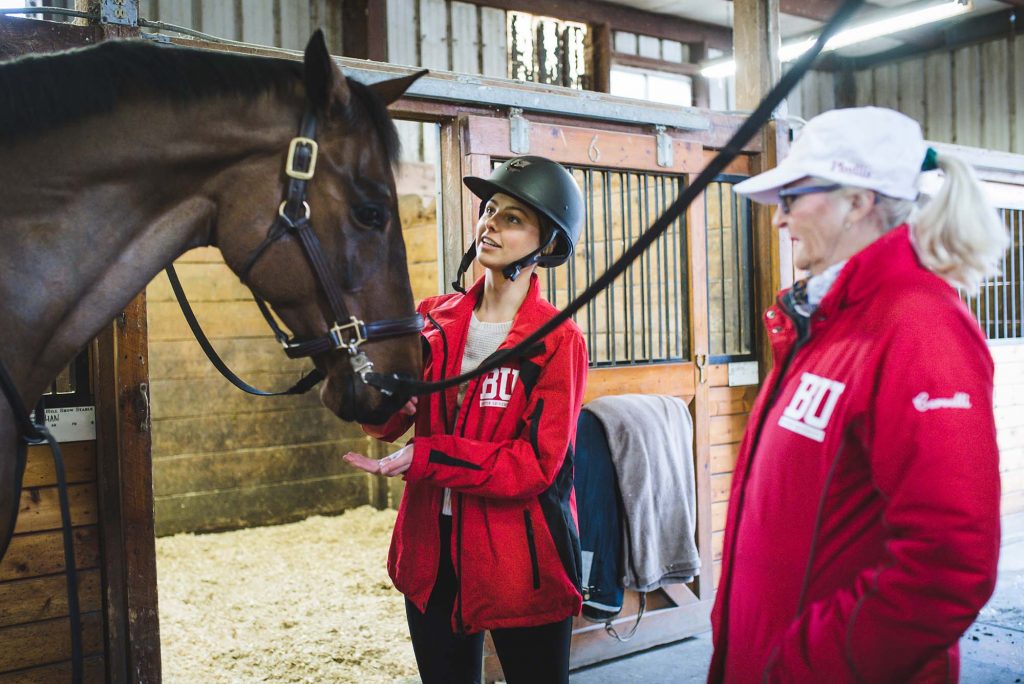 Photo of Lizzy Nevins, a young woman wearing a black riding helmet and red BU Equestrian jacket, standing next to BU Equestrian head coach Phyllis Cervelli, wearing a similar red jacket and white cap, in a barn barn. Lizzy is shown petting the head of a large dark brown horse with a black mane as Cervelli looks on to her right.