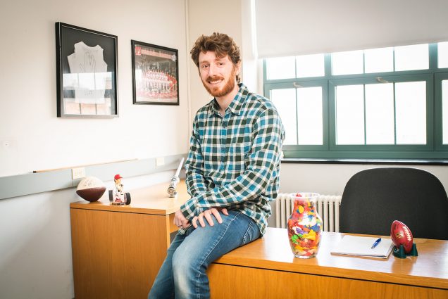 Photo of Marc Kornblueth, a Boston University College of Arts & Sciences post doc researcher, in his office. A white man with reddish brown short curly hair and short beard, sits on the edge of his desk. He wears a green and white plaid shirt and faded blue jeans as he poses and smiles for the camera.