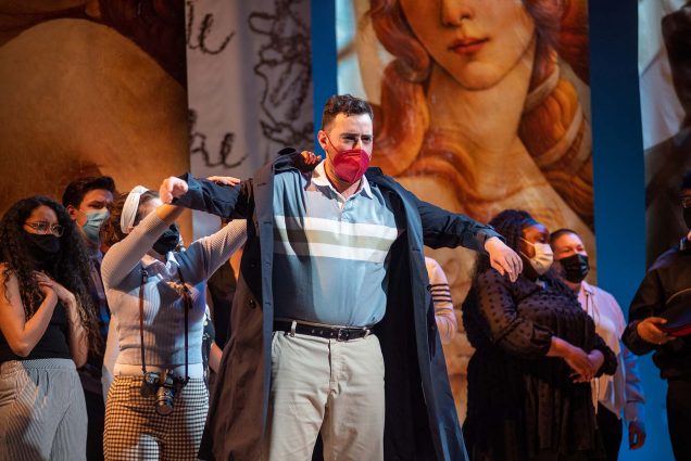 Photo of Yunus Akbas playing Guglielmo as he rehearses the Opera Institute’s Cosi fan tutte at the Tsai Performing Arts Center. On stage, a man with a stern facial expression and wearing a red face mask stands with arms splayed out to his sides and a person behind him helps hi put on a black jacket. Various people wearing masks behind him look out to the crowd with questioning and gossiping looks on their faces.