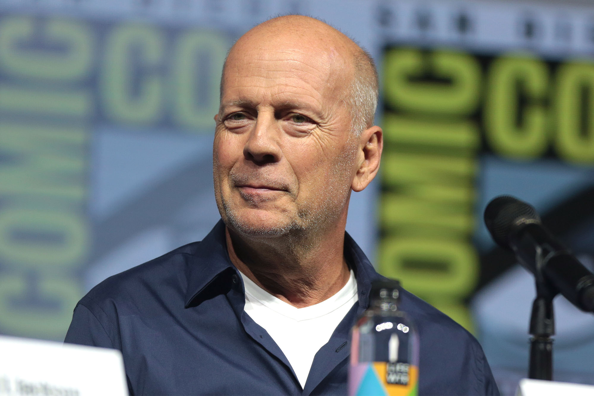 Bruce Willis at a speaking engagement wearing a blue collared shirt with a white undershirt