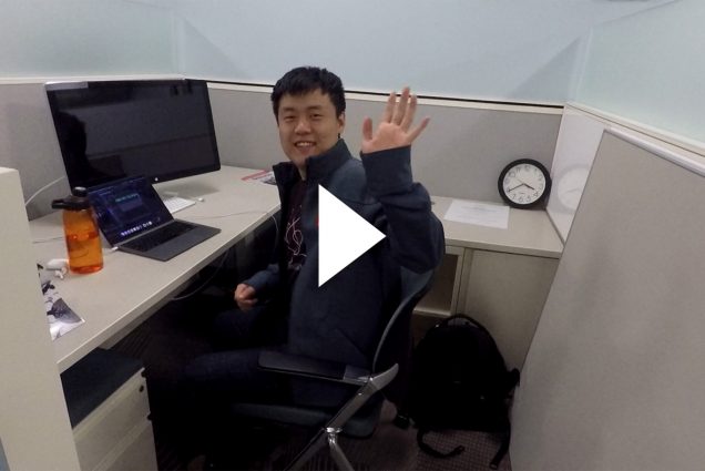Photo of Warren Liu, a young Asian man, waving tot he camera as he sits at a desk in a cubicle. An open laptop and Apple monitor sit in front of him. A white triangle video play button is overlaid over the image.