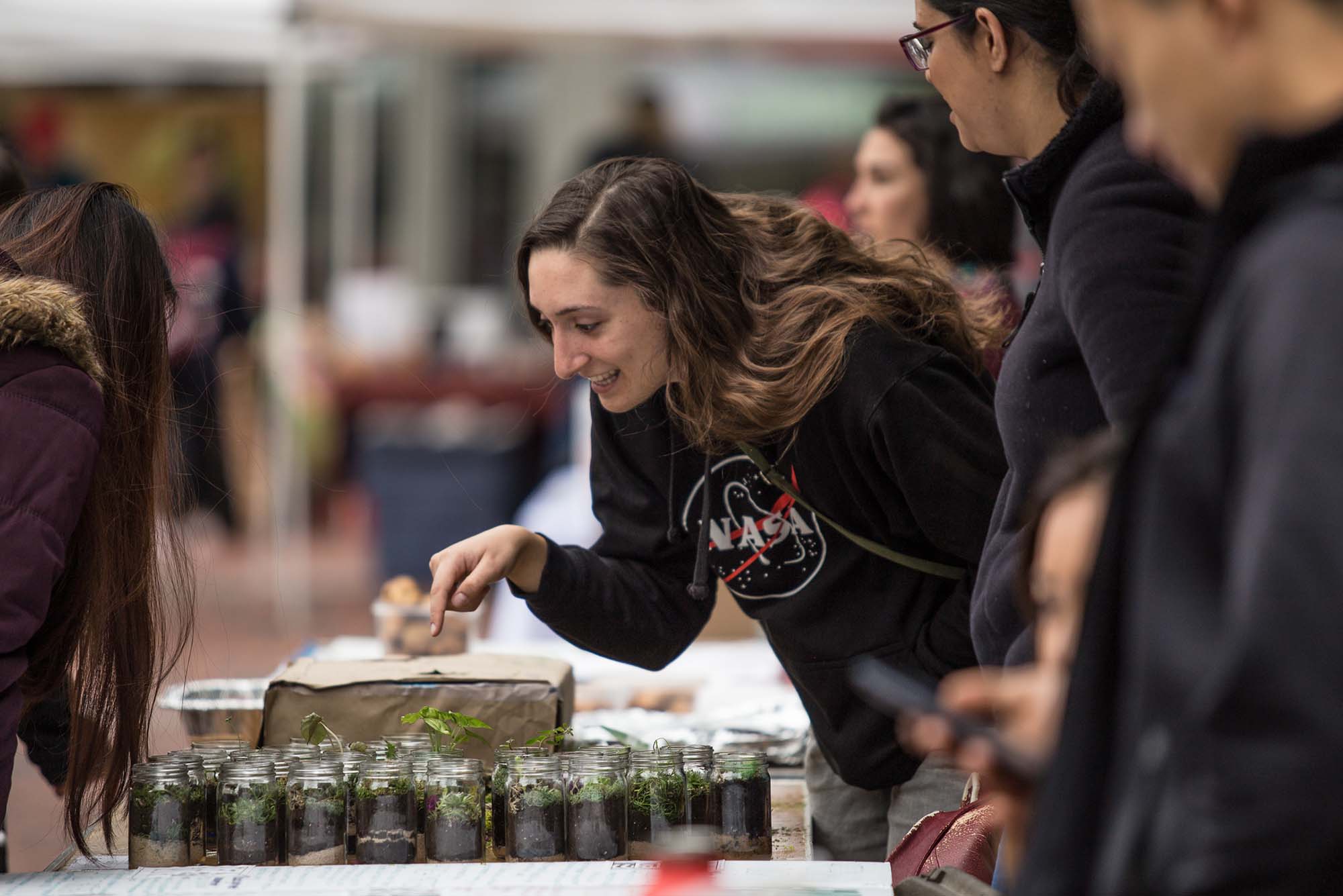 Photo of Tova Levin (CAS’18), a white woman with long light brown hair who wears a black NASA sweatshirt points at plants in mason jars on the table below her and smiles during the 2017 Earth Day Festival in the GSU Plaza. Other students looking at plants can be seen around her.