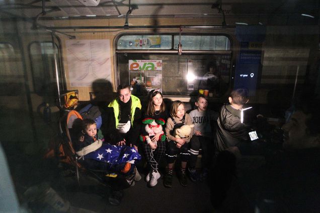 Photo of children, of various ages, sitting in a train car in a metro station that serves as a bomb shelter. They have blankets and some hold stuffed animals. Above them you can see subway advertisements. The photo was taken through a subway car window.