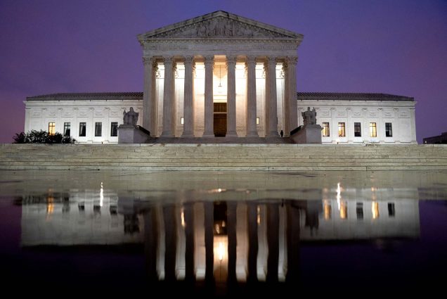 Photo of the facade of the supreme court at dusk. The white, Grecian facade is reflected in a puddle in front of the main steps. The sky is blueish purple and lights shine from behind the columns.