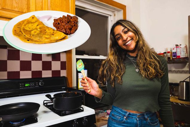 Photo of Neytra, a young Indian American woman with light brown skin and long brown, highlighted hair. She wears a green sweater and holds a spatula in her right hand. Behind her, you can see two black pots on a stove and a kitchen backsplash. At the top left of the photo is an image of the dish she cooked: curry made with red kidney beans and pureed tomatoes and flavored with spices like cloves, tumeric, and cinnamon with a side of roti. White play button is overlaid.