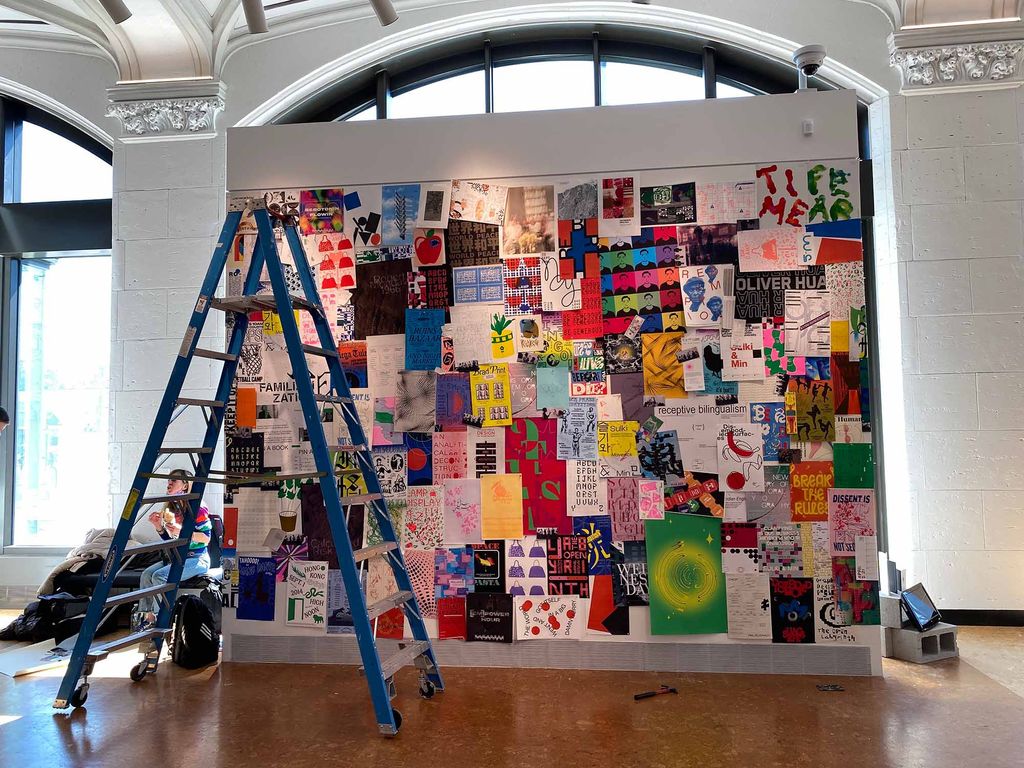 Photo of a large wall in Stone Gallery plastered with a series of posters from graphic design students. Papers vary in size, from postcard size to 12 by 24 inches, and have all kinds of colorful design, text, and images. A blue ladder is seen front left.