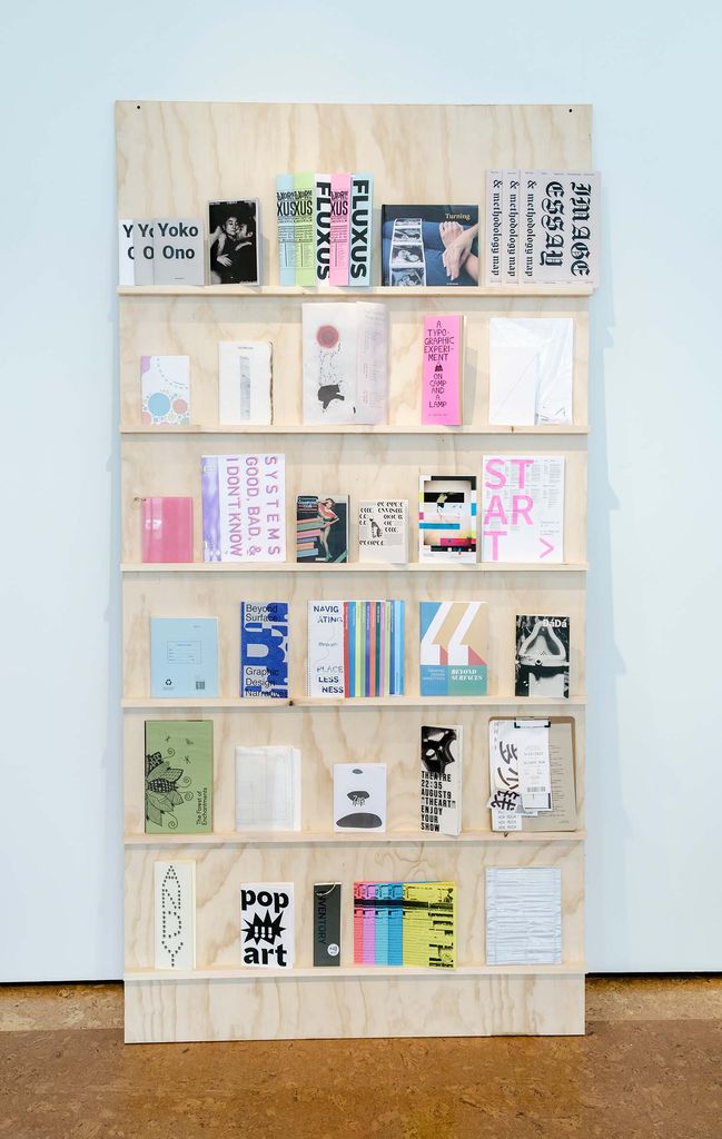 Photo of one of the installations from the MFA graphic design thesis show: an unfinished plywood bookshelf with zines of various sizes from students. Some titles include: "Yoko Ono, Pop Art, ANDY, Systems Good, Bad & I Don't Know"