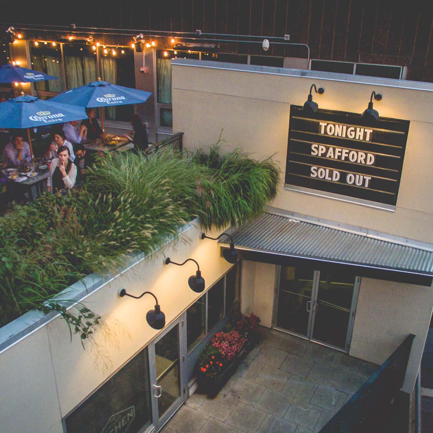 Photo of the Sinclair roofdeck. The marquee on the tan building reads "Tonight Spafford Sold out." To the left, patrons can be seen dining outside at tables with blue Corona umbrellas next to a railing with plants.