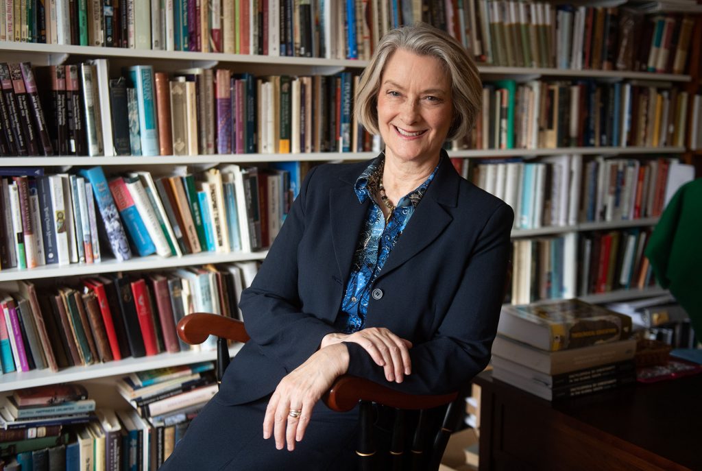 Photo of Professor  Dana Robert of STH. She is a middle-aged White woman with short salt and pepper hair. She wears a black blazer and blue blouse, and smiles as she sits in a wooden chair, her hands crossed in her lap. Behind her, a large white bookshelf filled with books is seen.