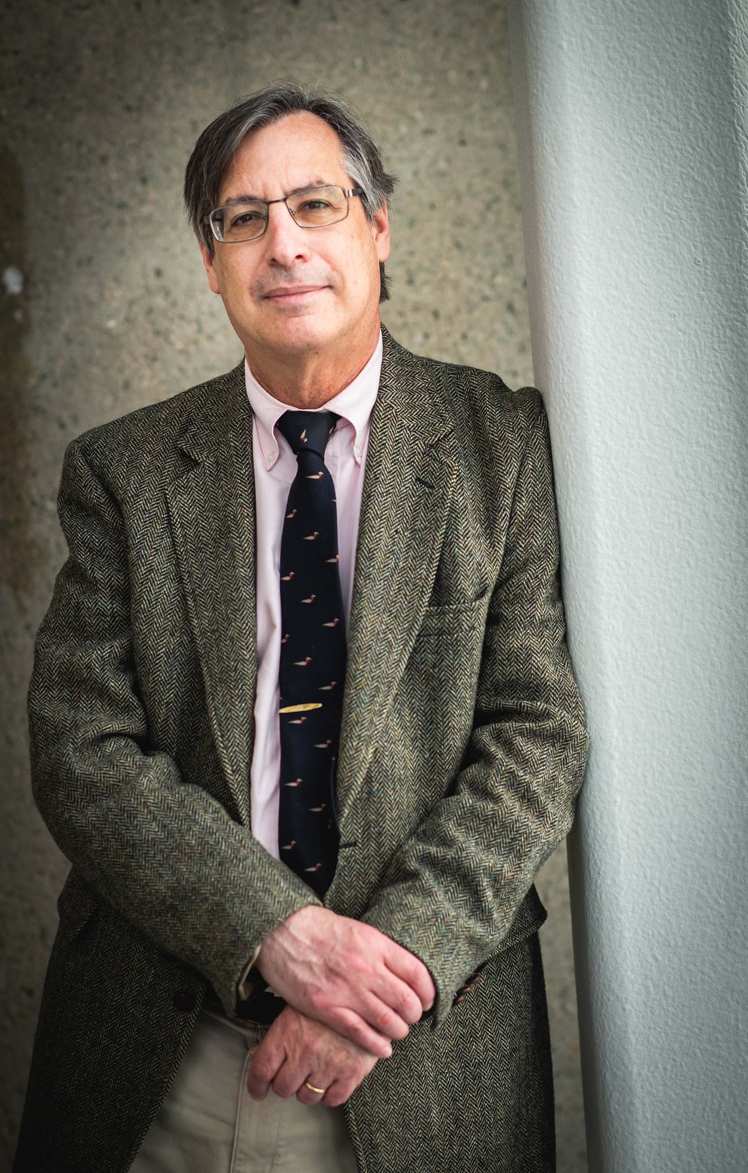 Photo of Gary Lawson, School of Law professor of Law. He is a middle-aged white man with glasses and salt and pepper hair. He wears a speckled gray-green tweed suit, pink button down and black tie. He smiles slightly and holds his arm with his hand and learns against a white wall.