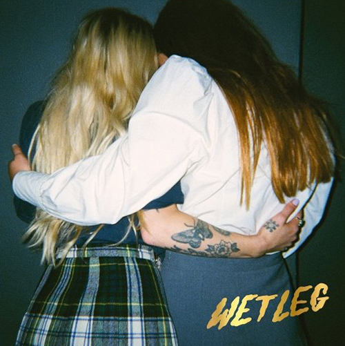 Album cover for Wet Leg's self-titled album, "Wet Leg". Cover is a photo of the back of two people with long hair. On the left a blonde person stands with a plaid skirt and top and on the right a brunette stands with a white long-sleeved dress shirt and a sweatshirt wrapped around their waist that reads "Wet Leg". Both have an arm around each other and are huddled close together.