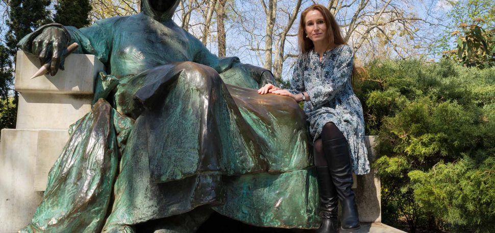 Photo of Theodora Goss in Budapest park with teal rusting bronze statue of Anonymus, a well known landmark. A white and red hot air balloon can be seen in the distance of the background, behind naked tree branches.