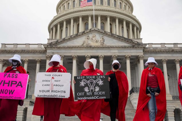 Photo of abortion-rights protesters dressed in costumes from the "Handmaid's Tale," holding signs in front of the U.S. Capitol building during a demonstration in Washington, DC. Four masked women stand with long red robes and white bonnets. They hold various signs protesting an abortion ban. One prominent sign reads "Keep your laws off my body, handmaidsDC.com".